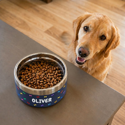 Golden retriever waiting beside a navy Personalised Pattern Dog Bowl on a table, the name OLIVER set into a band of rainbow-coloured bones