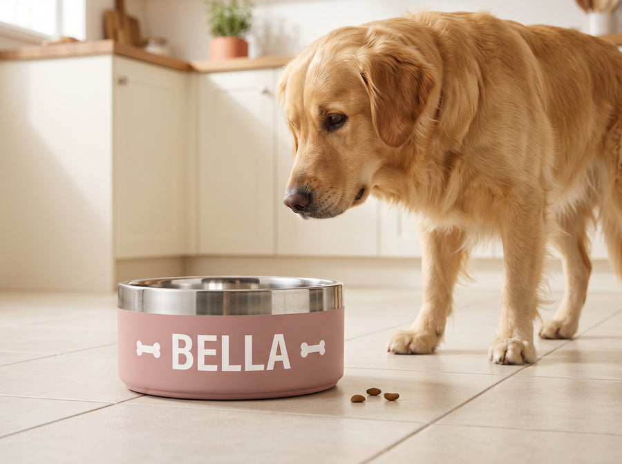 A golden retriever beside a personalised pink dog bowl printed with BELLA on a tiled kitchen floor.