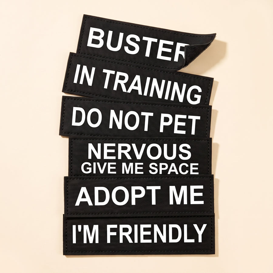 Six removable velcro patches for a personalised dog harness fanned out on a cream background, reading BUSTER, IN TRAINING, DO NOT PET, NERVOUS GIVE ME SPACE, ADOPT ME and I'M FRIENDLY.