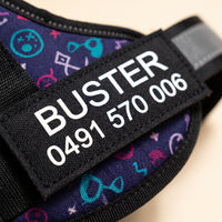 Close crop of the removable name patch on a personalised dog harness, printed BUSTER and a mobile number in white on black.